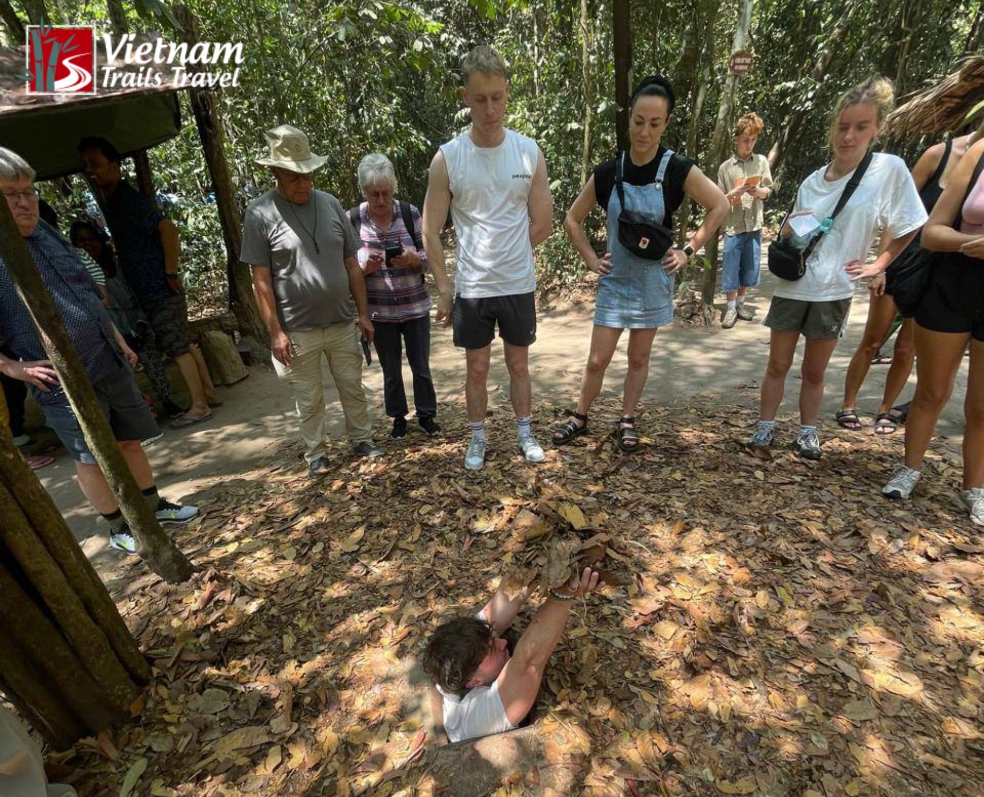 A group of international tourists watching a man demonstrate how to enter a tiny, camouflaged trapdoor hidden under dry leaves at the Cu Chi Tunnels