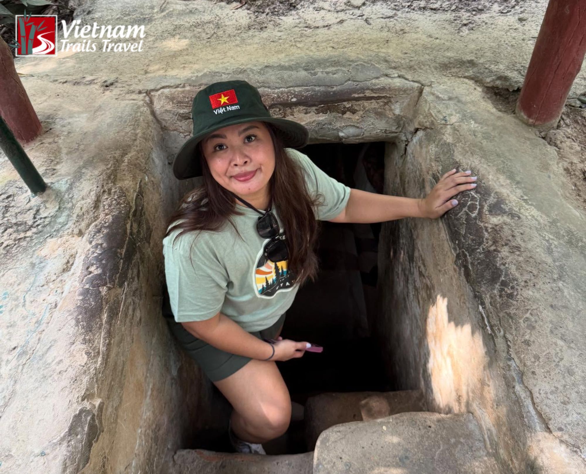 A smiling female tourist wearing a Vietnam flag bucket hat, exploring the narrow concrete entrance of the Cu Chi Tunnels