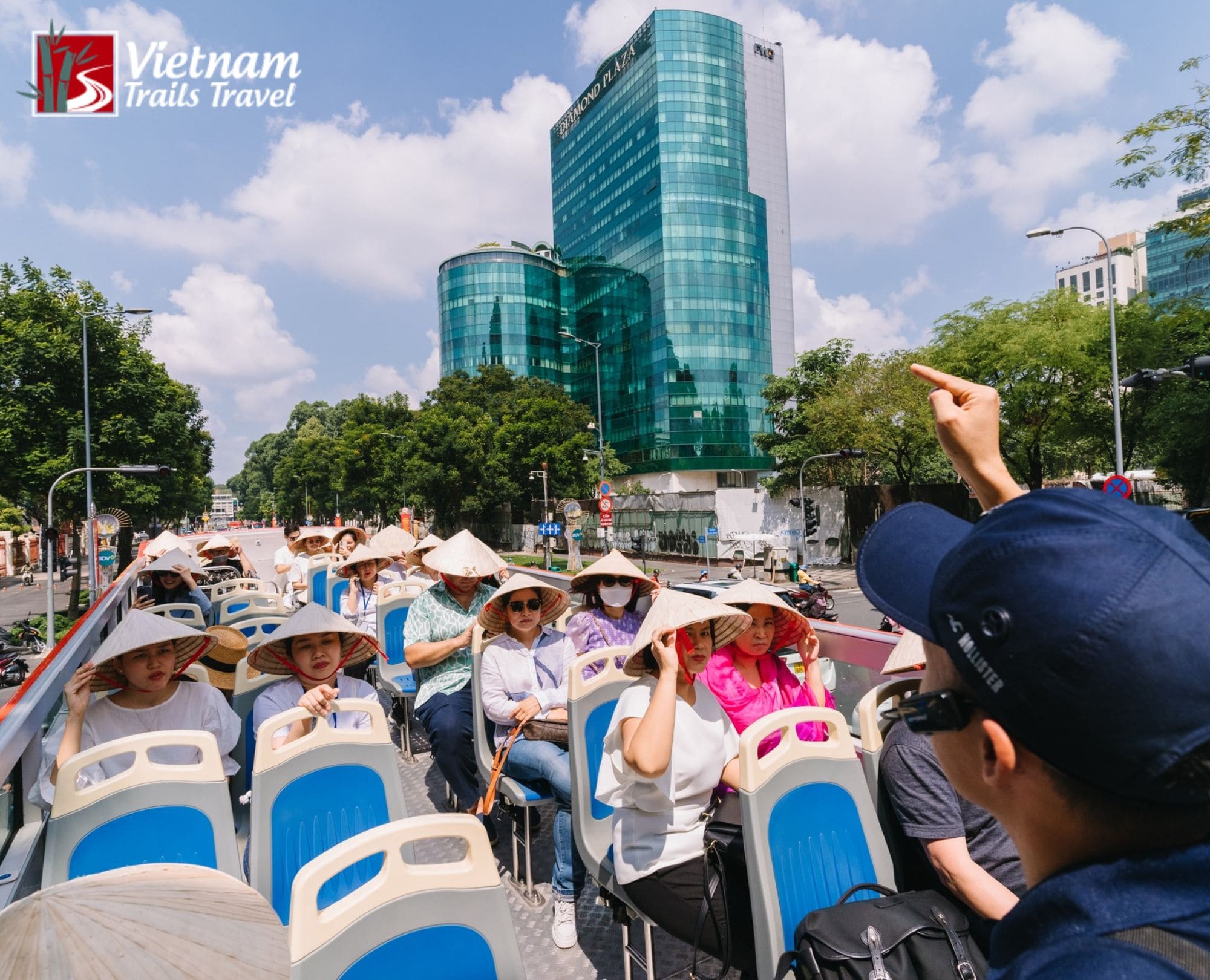 Tourists wearing traditional Vietnamese conical hats (non la) sit on the open deck of a double-decker sightseeing bus in Ho Chi Minh City