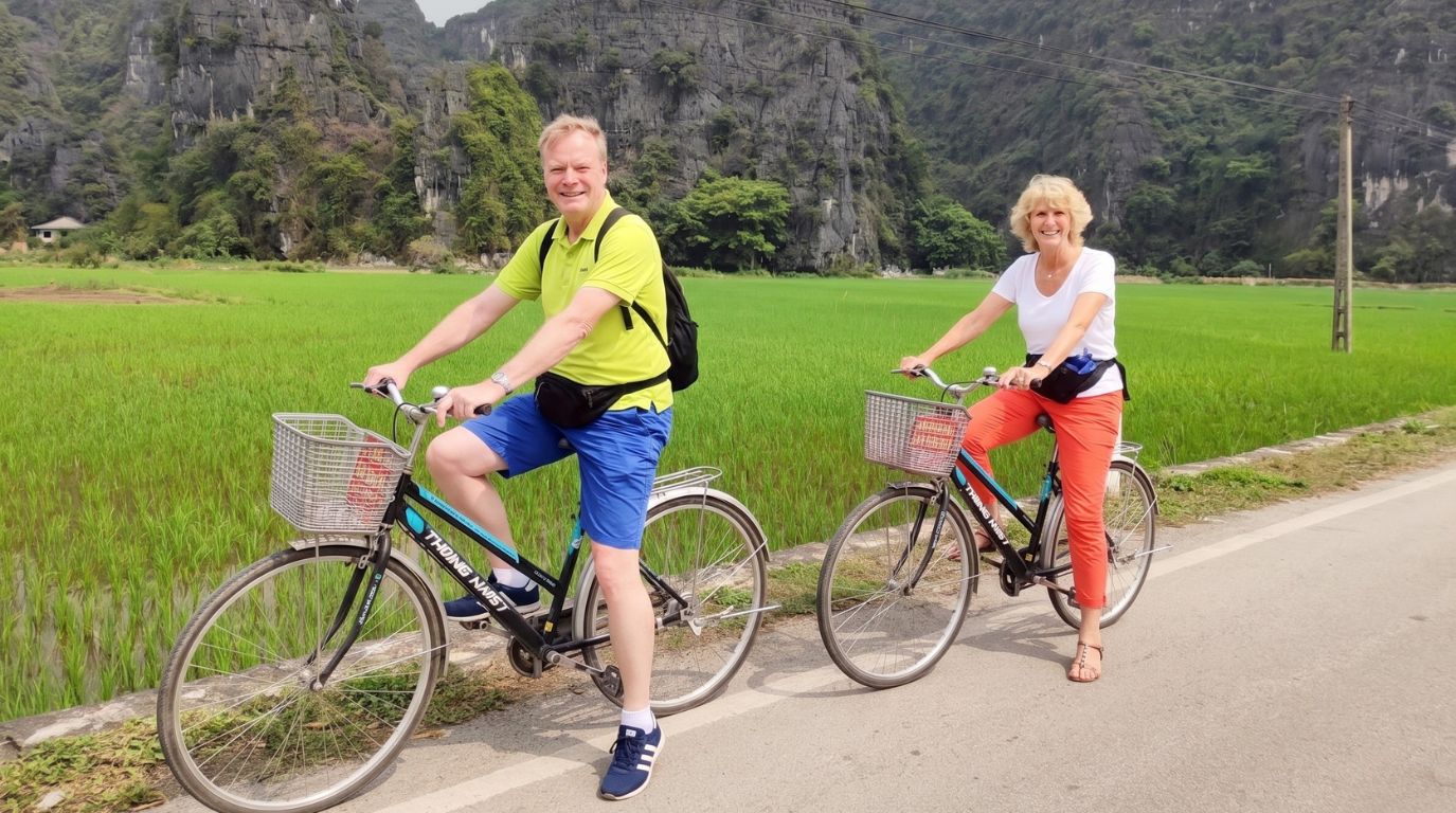 A couple cycling along a scenic country road next to lush green rice paddies and limestone karsts in Ninh Binh