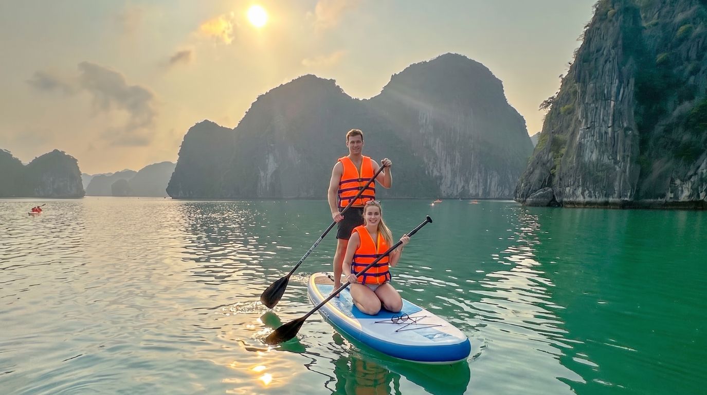 A couple wearing orange life jackets on a single stand-up paddleboard at sunset, surrounded by towering limestone islands in Lan Ha Bay