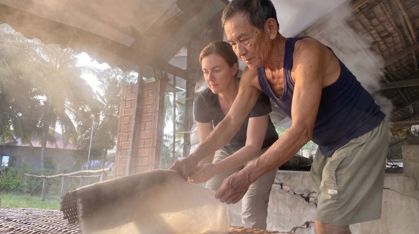 A female foreign tourist learning the traditional craft of making rice noodles with a local elder in a Mekong Delta village