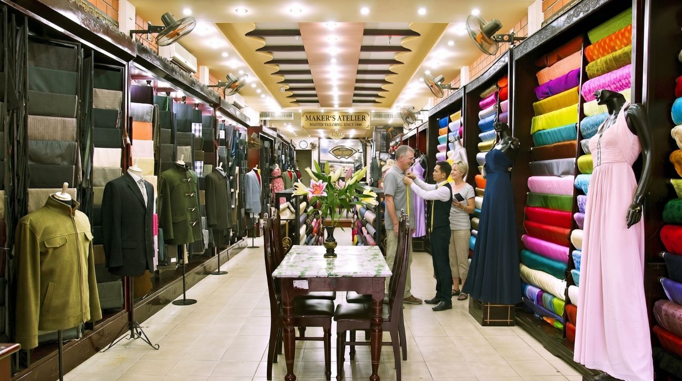 A male tourist getting measured by a local tailor inside a colorful custom clothing shop in Hoi An