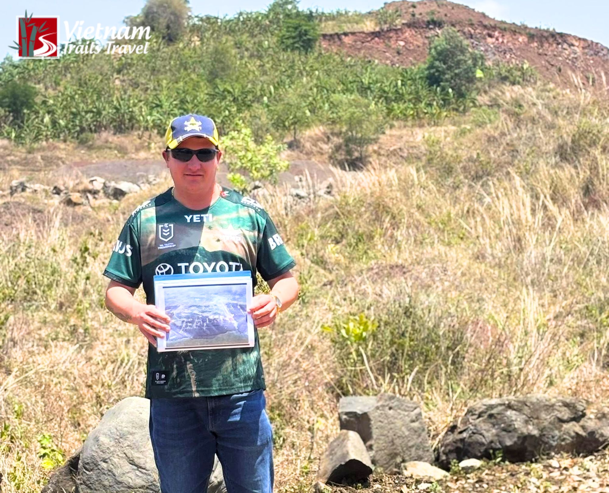 A man stands in a dry, grassy field at the base of SAS Hill and holding a laminated historical photo of the area