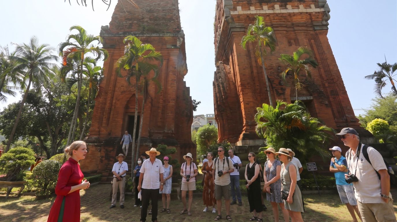 A tour guide in a red Ao Dai speaking to a group of international tourists in front of the ancient Cham Twin Towers