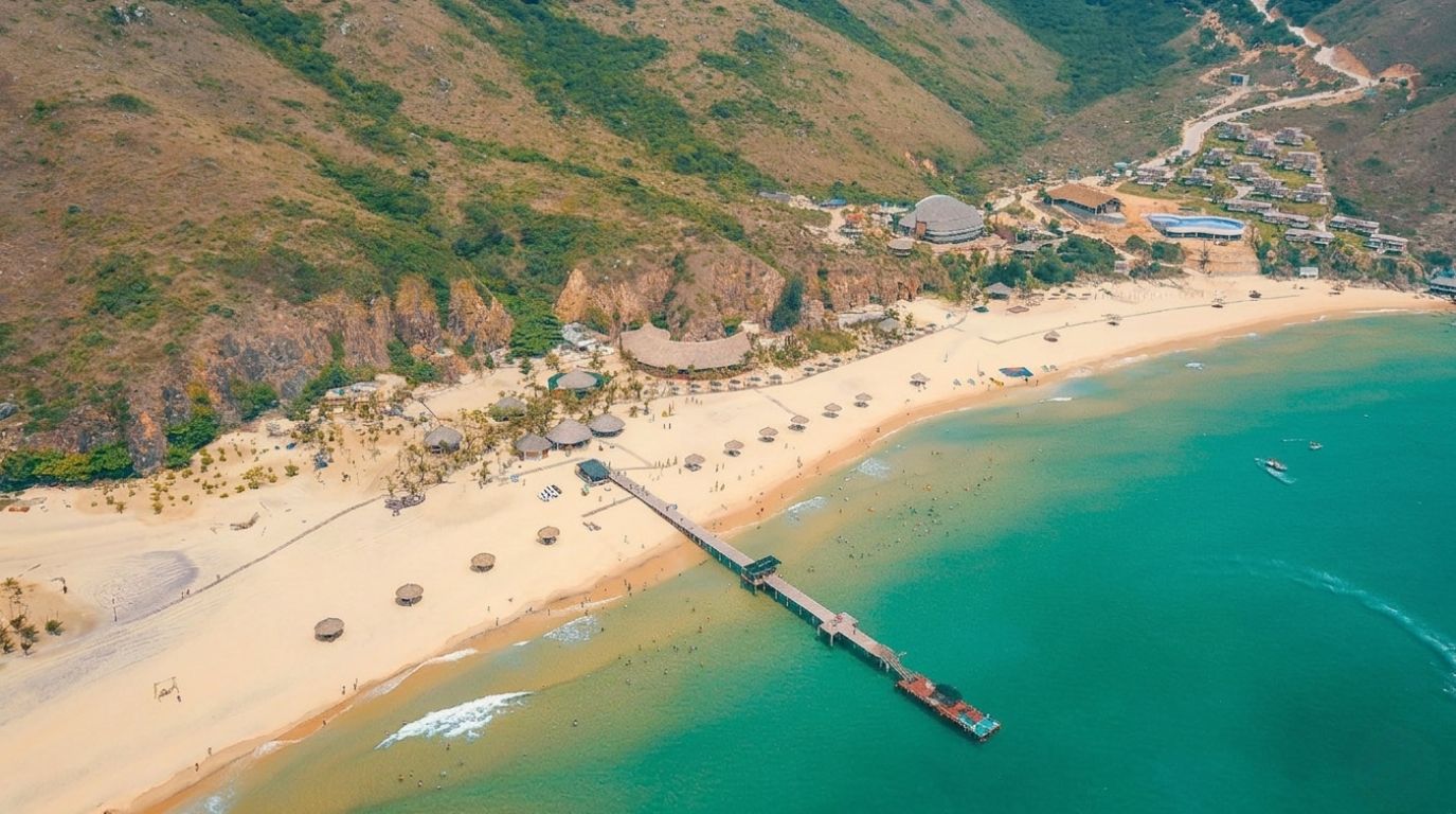 Aerial view of Ky Co beach with turquoise water, a long wooden pier, and resort huts nestled against green hills