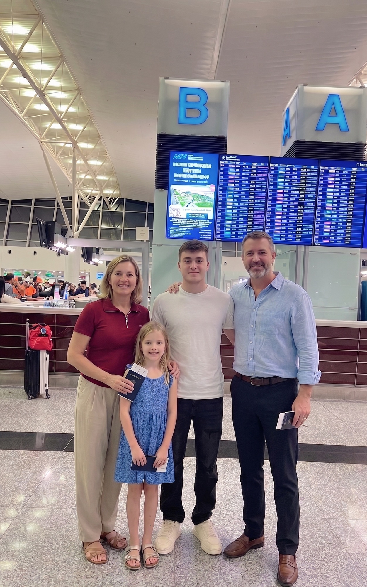 Happy foreign family of four holding passports and posing for a travel photo in front of the flight information screens at Noi Bai international airport terminal in Vietnam