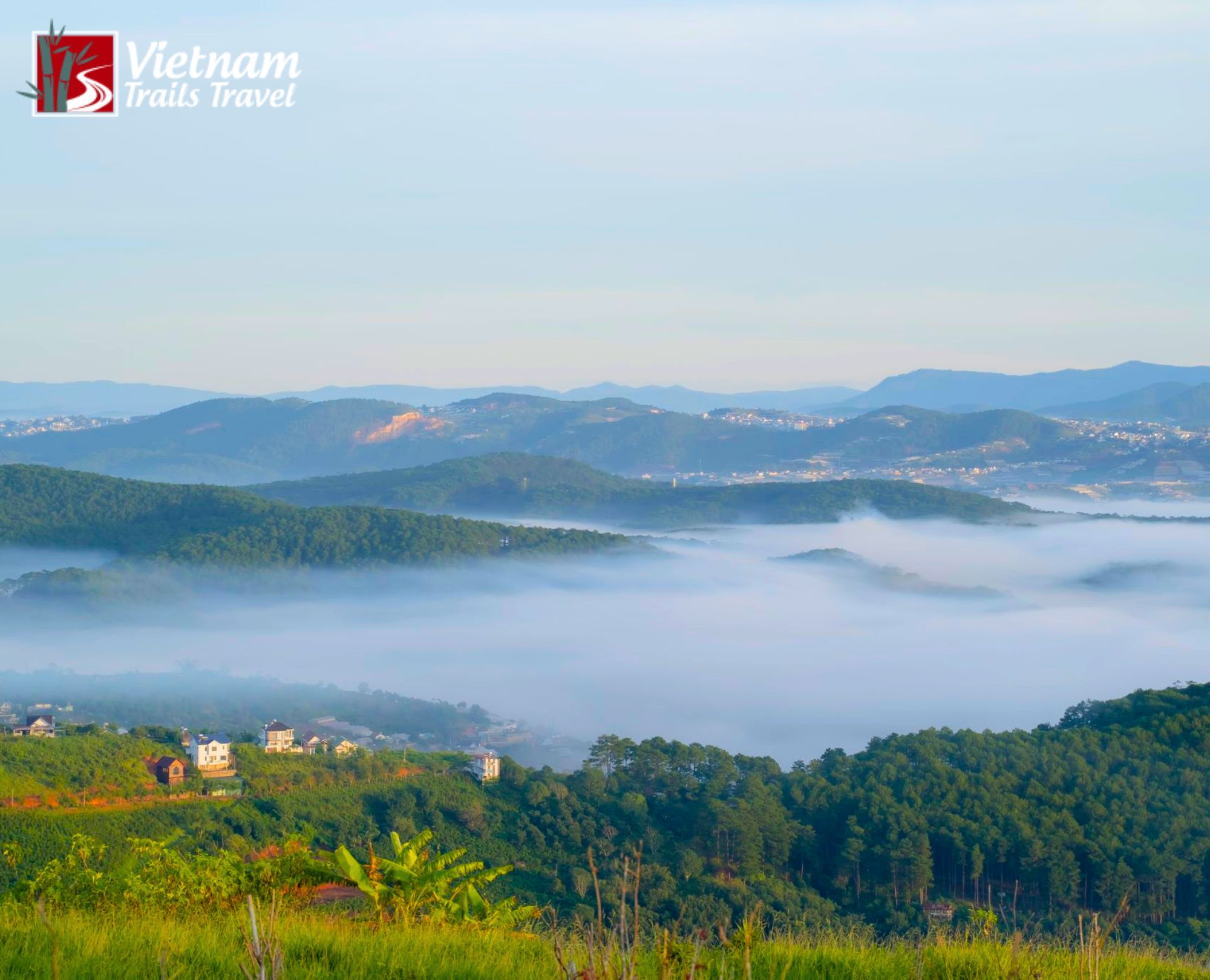 Misty rolling green hills and valleys dotted with houses, with fog blanketing the landscape and mountain ranges fading into the distance under a soft blue sky