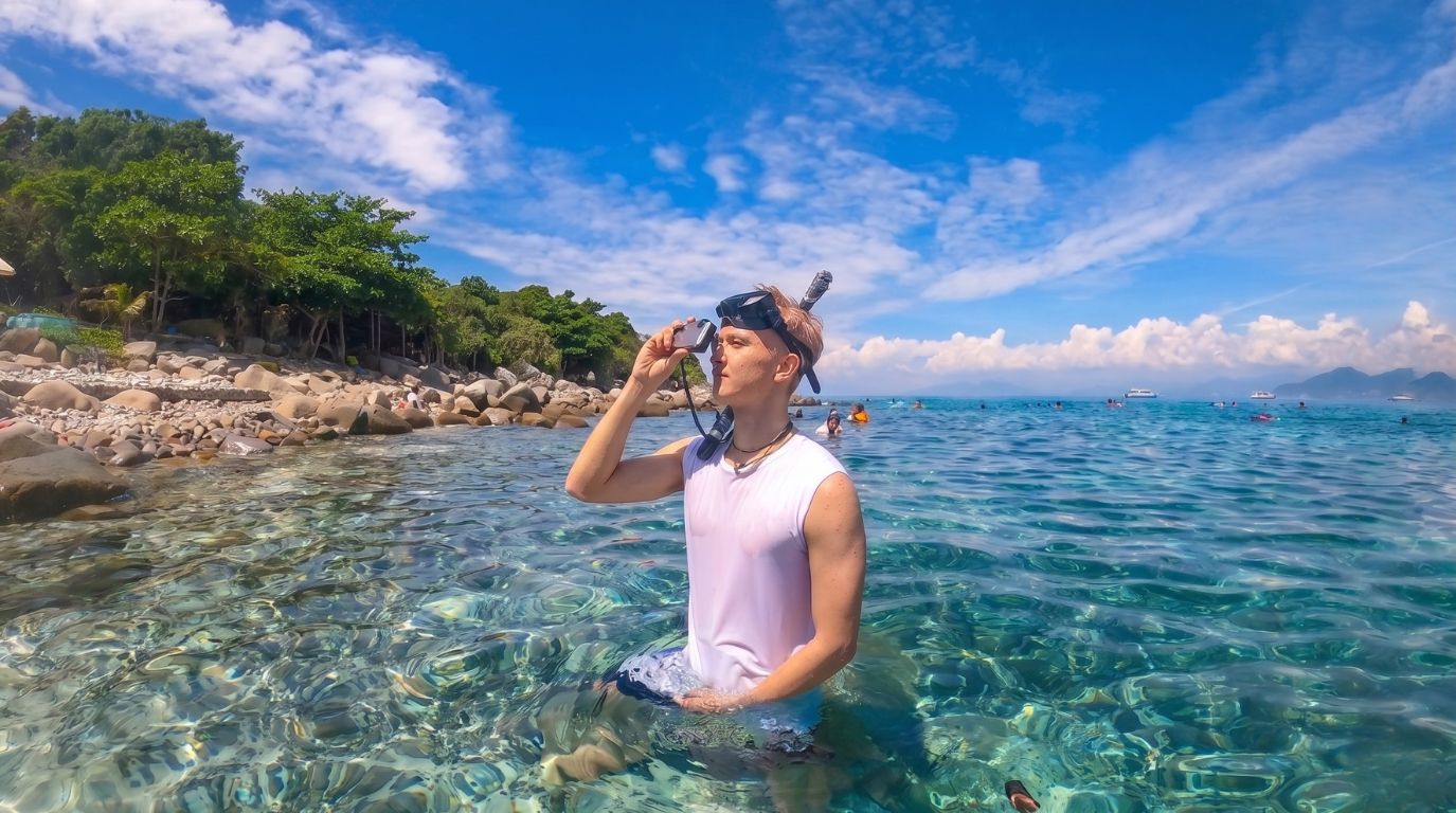 Tourist wearing snorkeling gear standing in the crystal clear blue waters near a rocky island in Hon Mun island