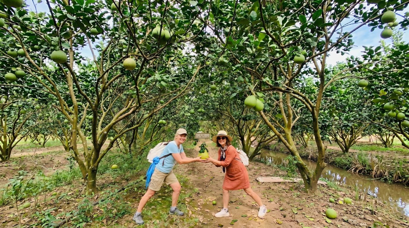 Two female tourists holding a fresh pomelo while exploring a lush green fruit orchard in Can Tho