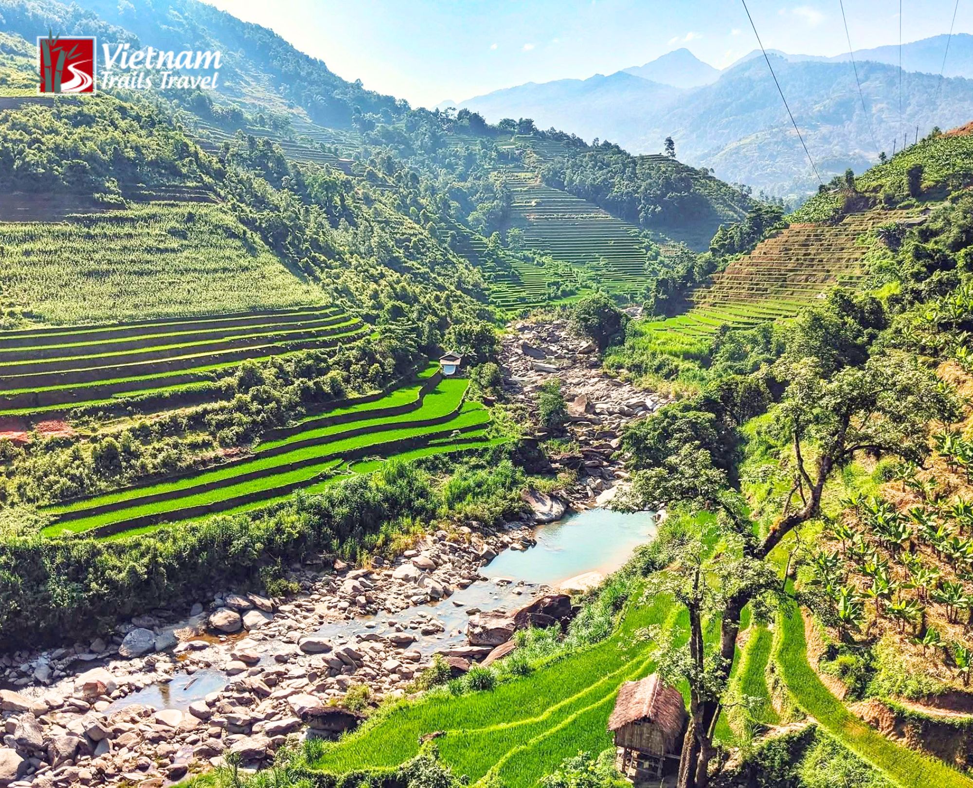 A river snakes through the lush Ho Thau valley, framed by vibrant green terraced paddies and thatched huts