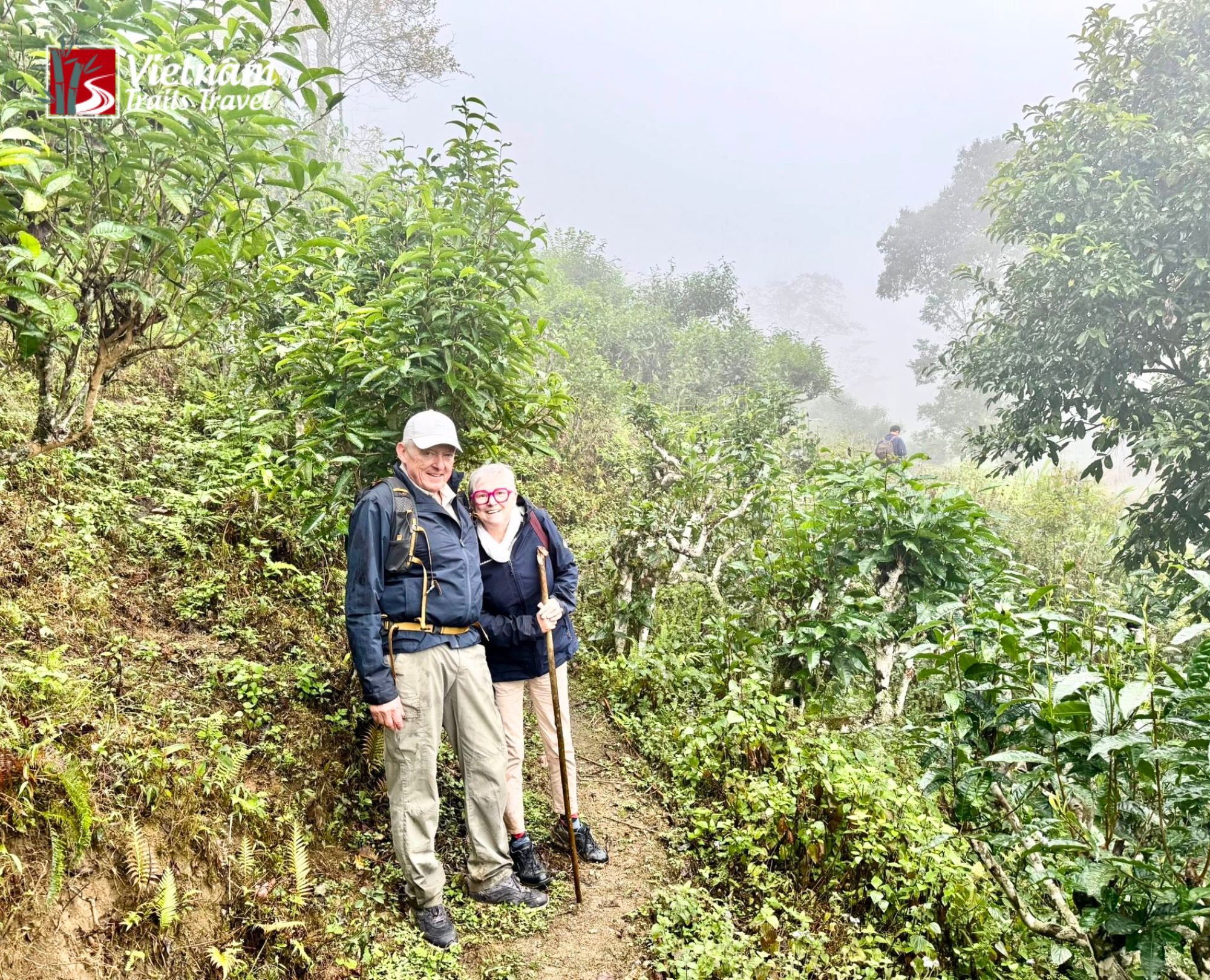 Two hikers trekking in Hoang Su Phi along a narrow dirt path surrounded by dense green foliage